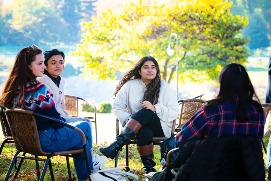 Four students seated in wicker chairs outdoors, talking amid autumn foliage.