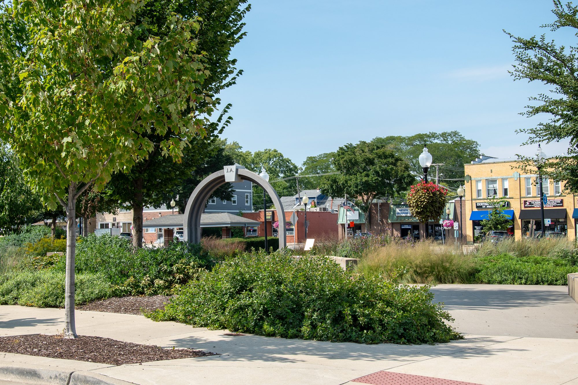 Landscaped downtown plaza with a metal arch sculpture and two-story commercial buildings.