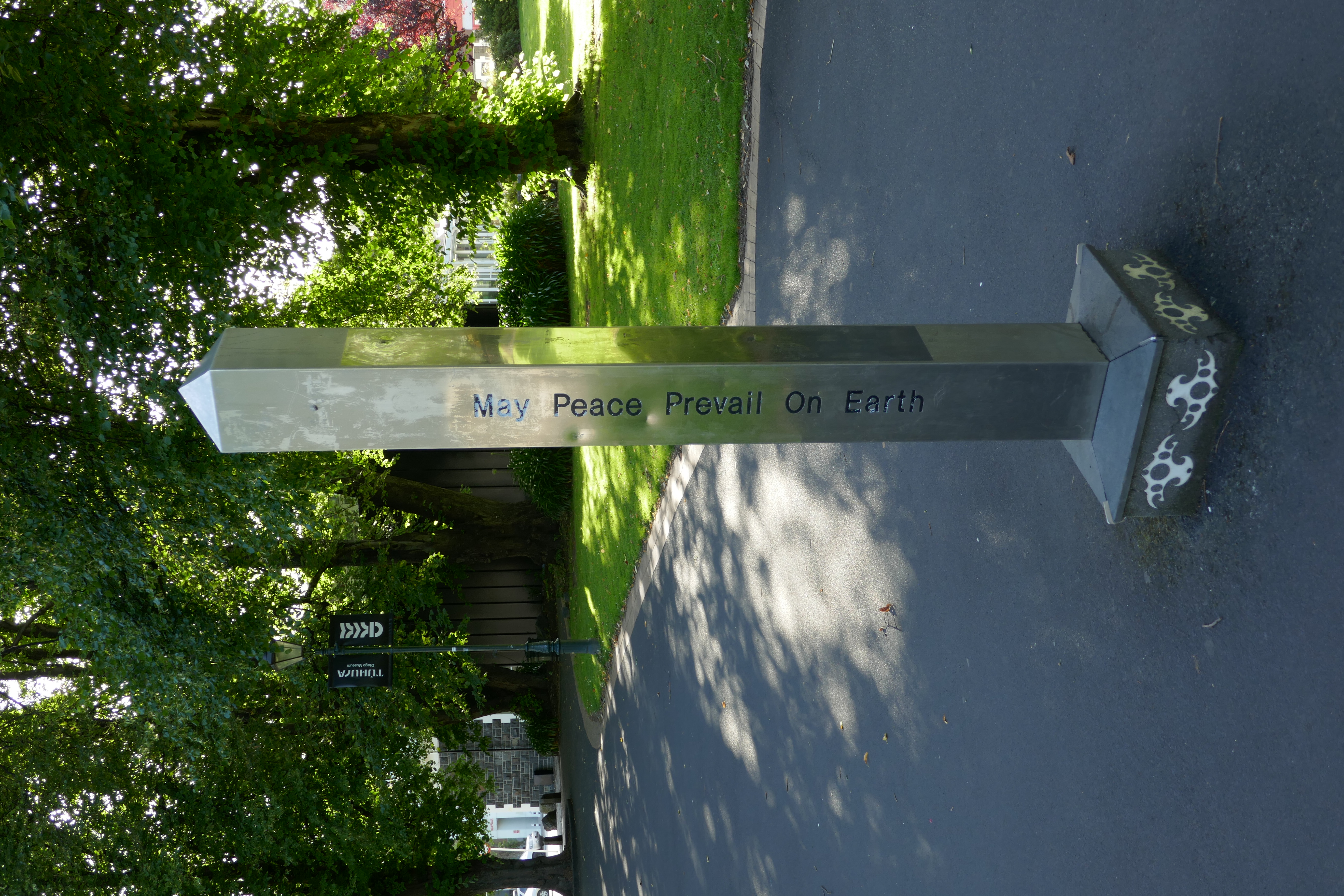 Tall metallic obelisk Peace Pole inscribed with 'May Peace Prevail on Earth' standing outdoors.