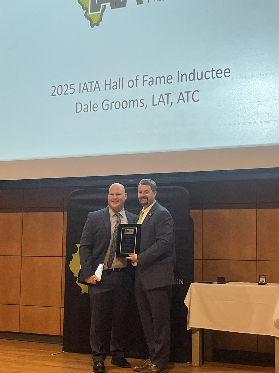 Two men in suits posing with a plaque at a Hall of Fame induction ceremony with a projection screen behind them.