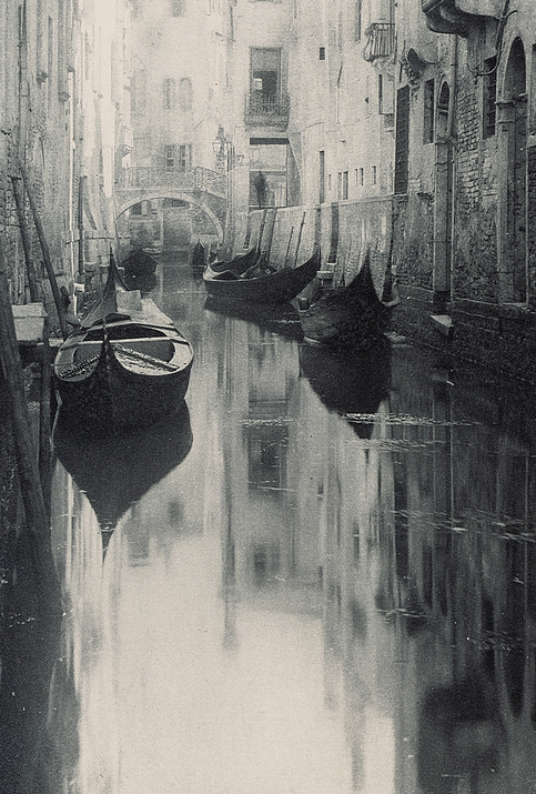 Black-and-white photograph of a narrow canal with gondolas and historic buildings.