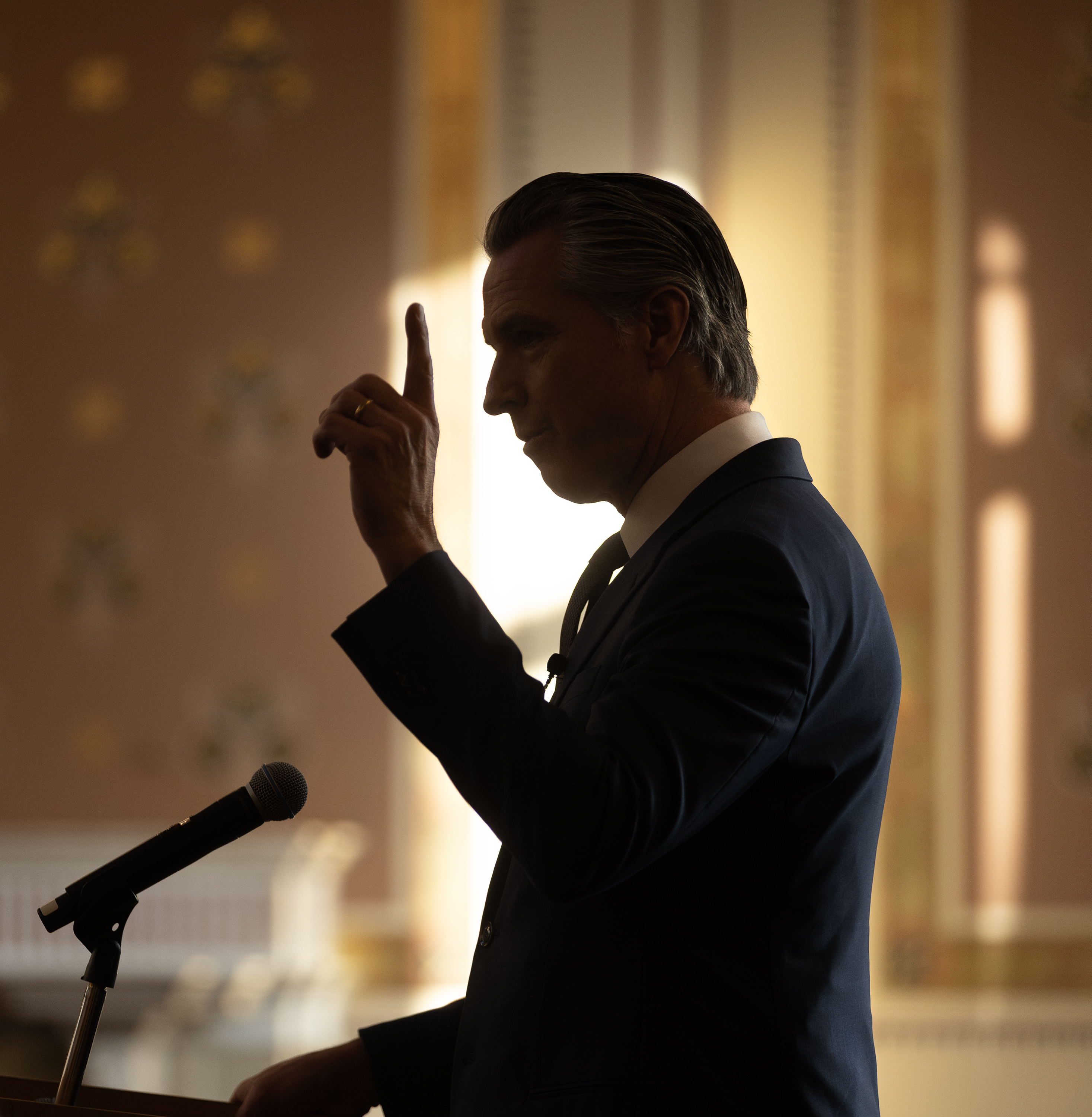 Man in a dark suit gestures at a podium, dramatically backlit against a golden background.