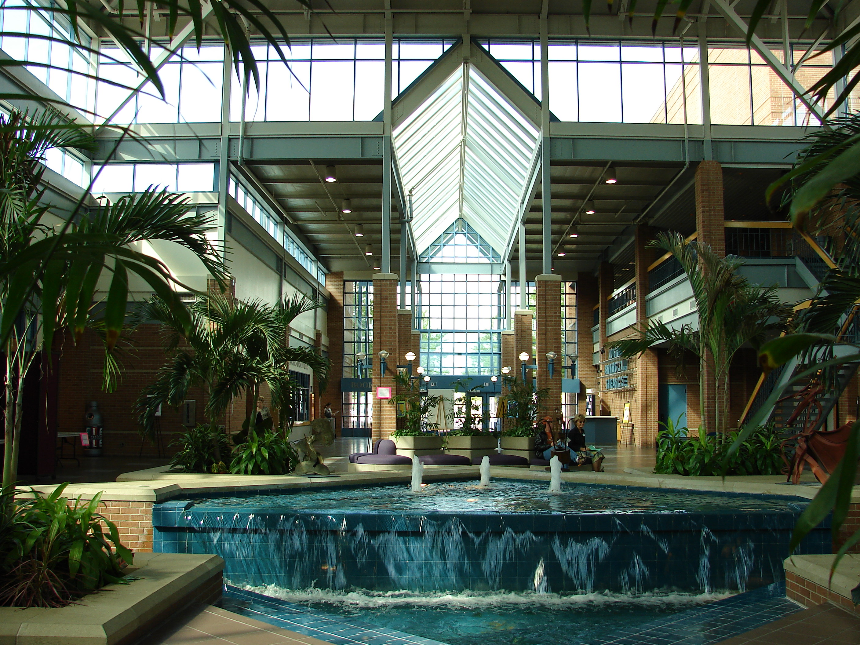 Sunlit atrium interior with tiered fountain, palm trees, brick columns, and glass skylight roof.