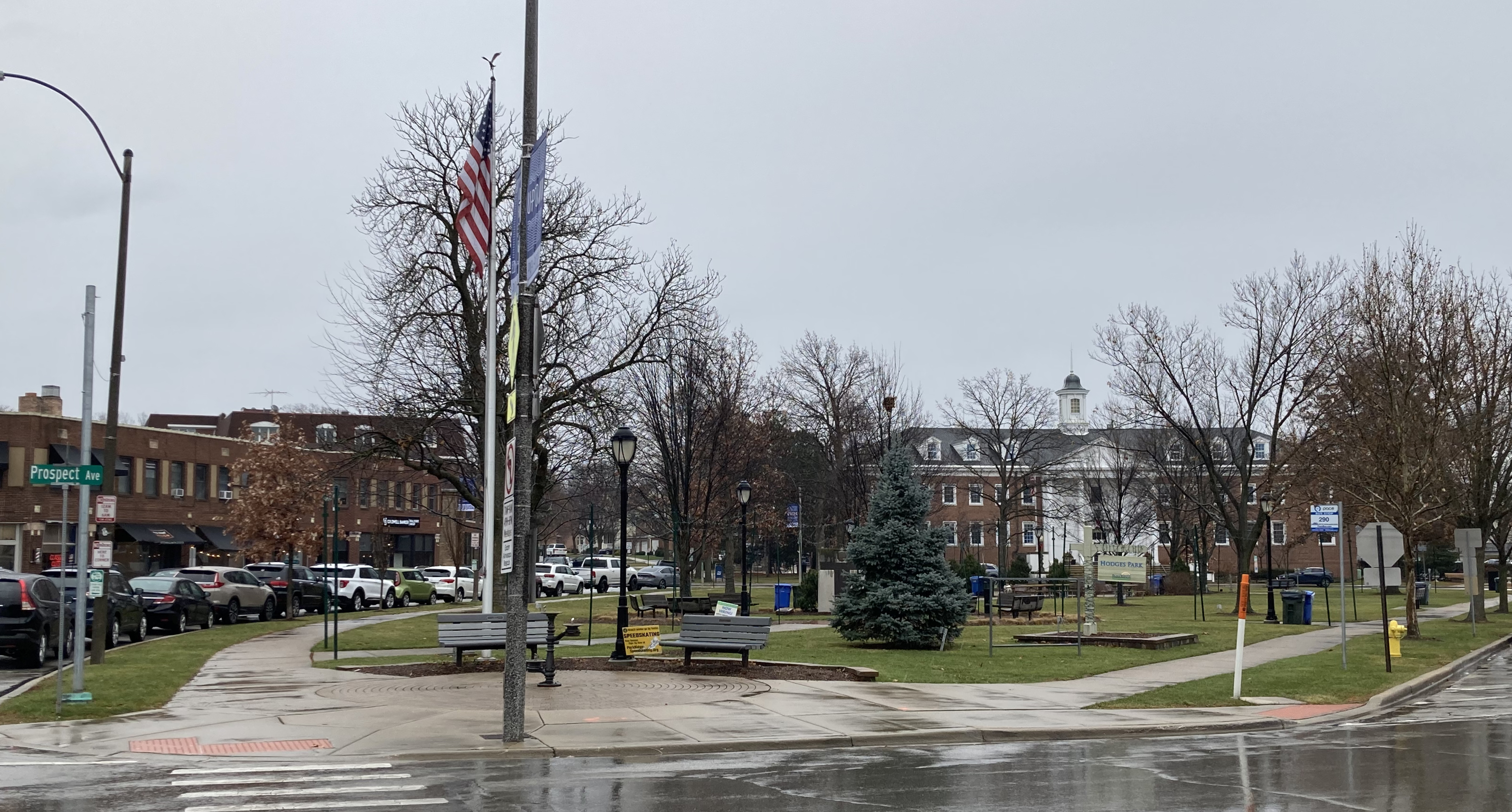 Park with bare trees, benches, an American flag, and a brick building in the background.