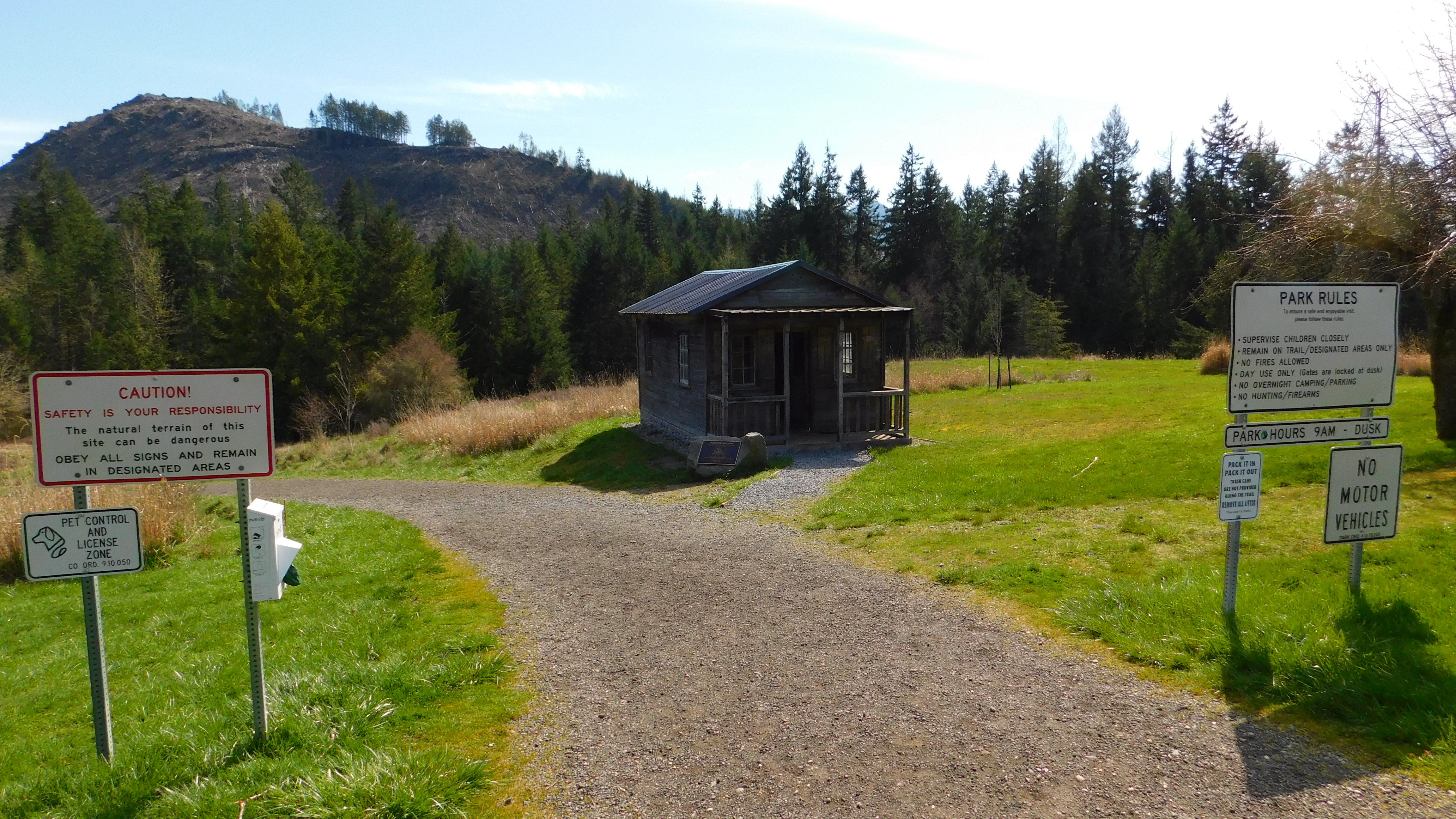 Gravel path leads to a wooden trailhead shelter with park signs and a rocky hillside.