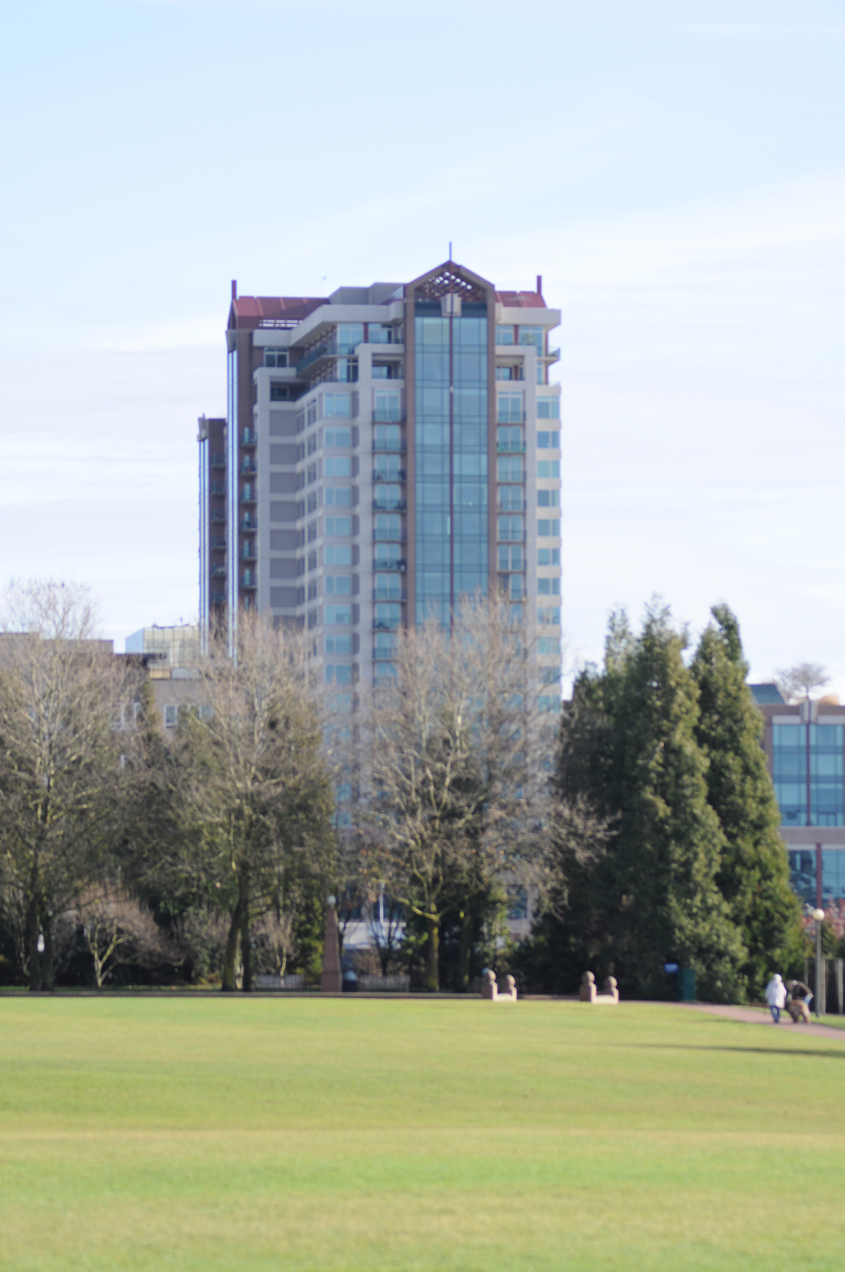 Tall glass office tower rising above a green park lawn with trees in the foreground.