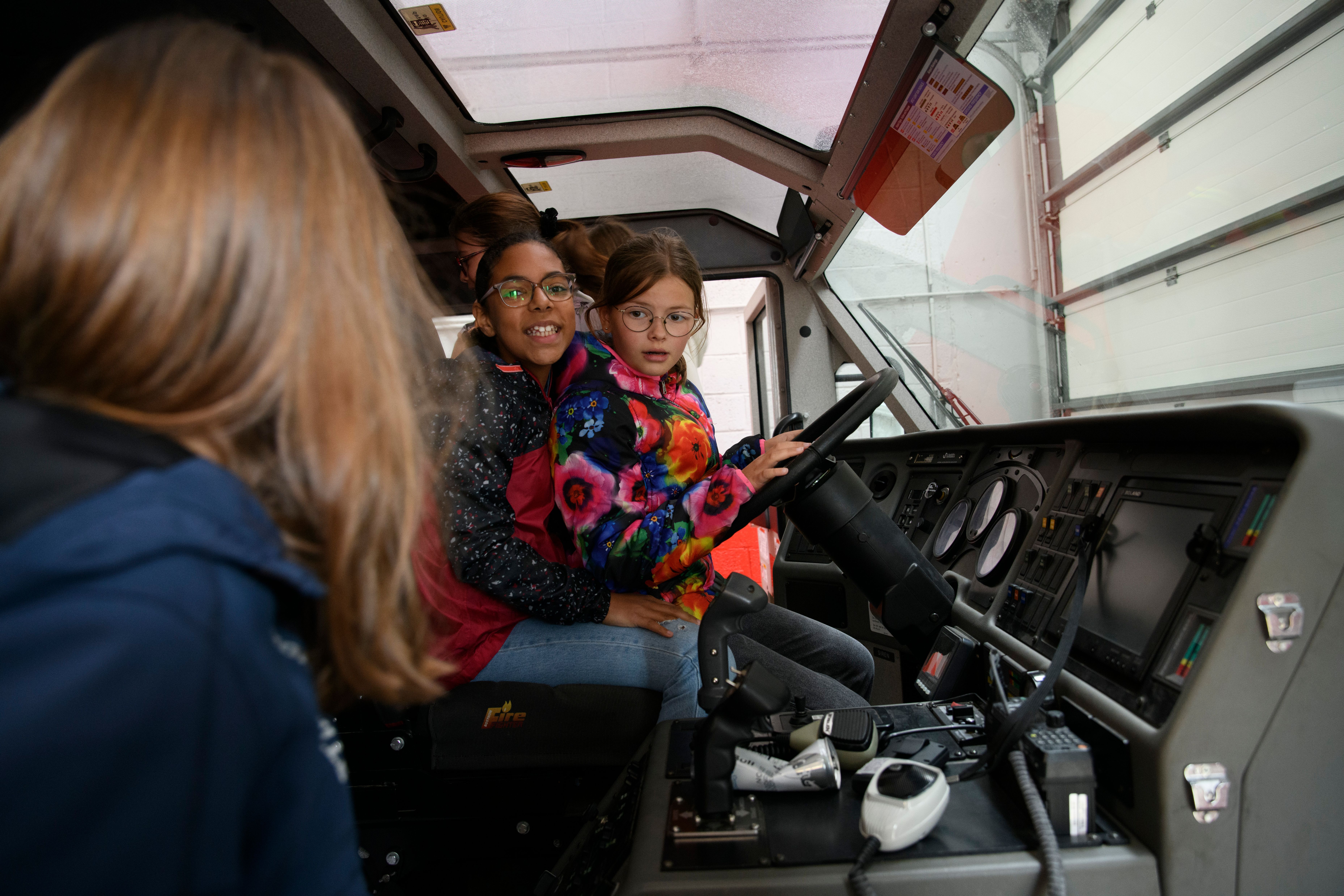 Two children sit in the cab of a large fire truck, one gripping the steering wheel.