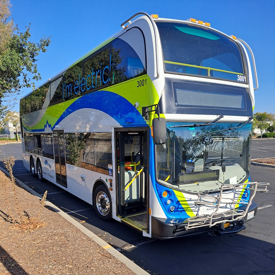 A green, blue, and white double-decker electric bus parked in a transit yard.