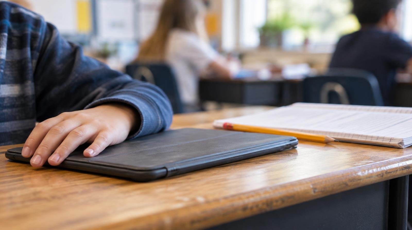 A closed tablet computer on a wooden desk next to a pencil and open notebook.