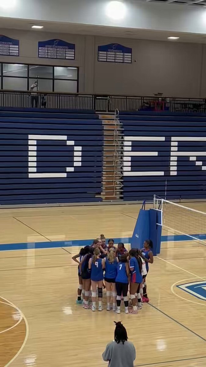 Volleyball players in blue and white uniforms huddle on a hardwood gym floor.