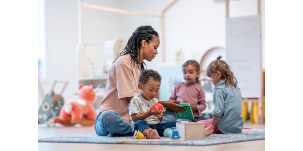 Adult sitting on a rug with three young children playing with colorful toys in a bright playroom.