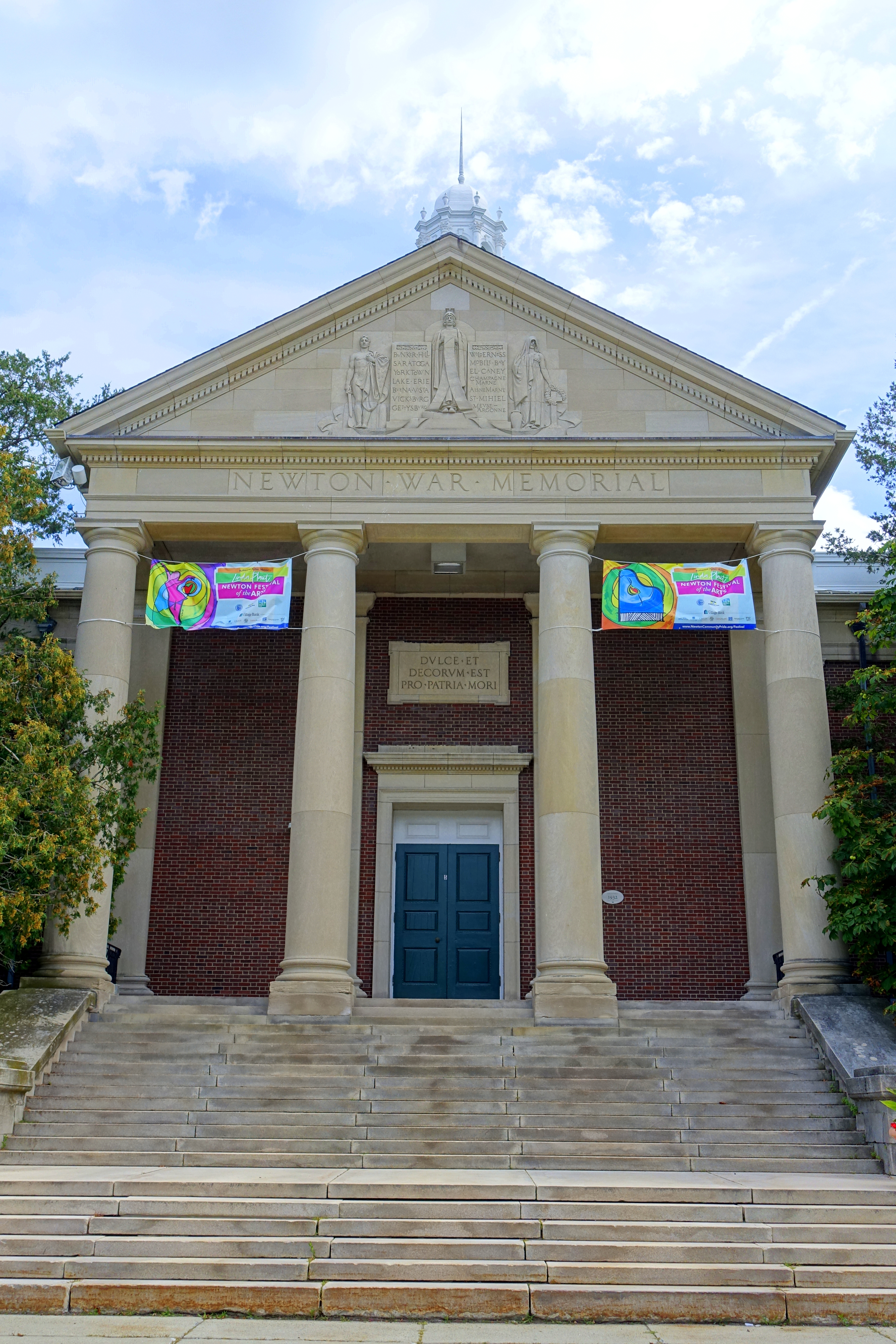 Neoclassical brick building entrance with columns, teal door, stone steps, and colorful banners.