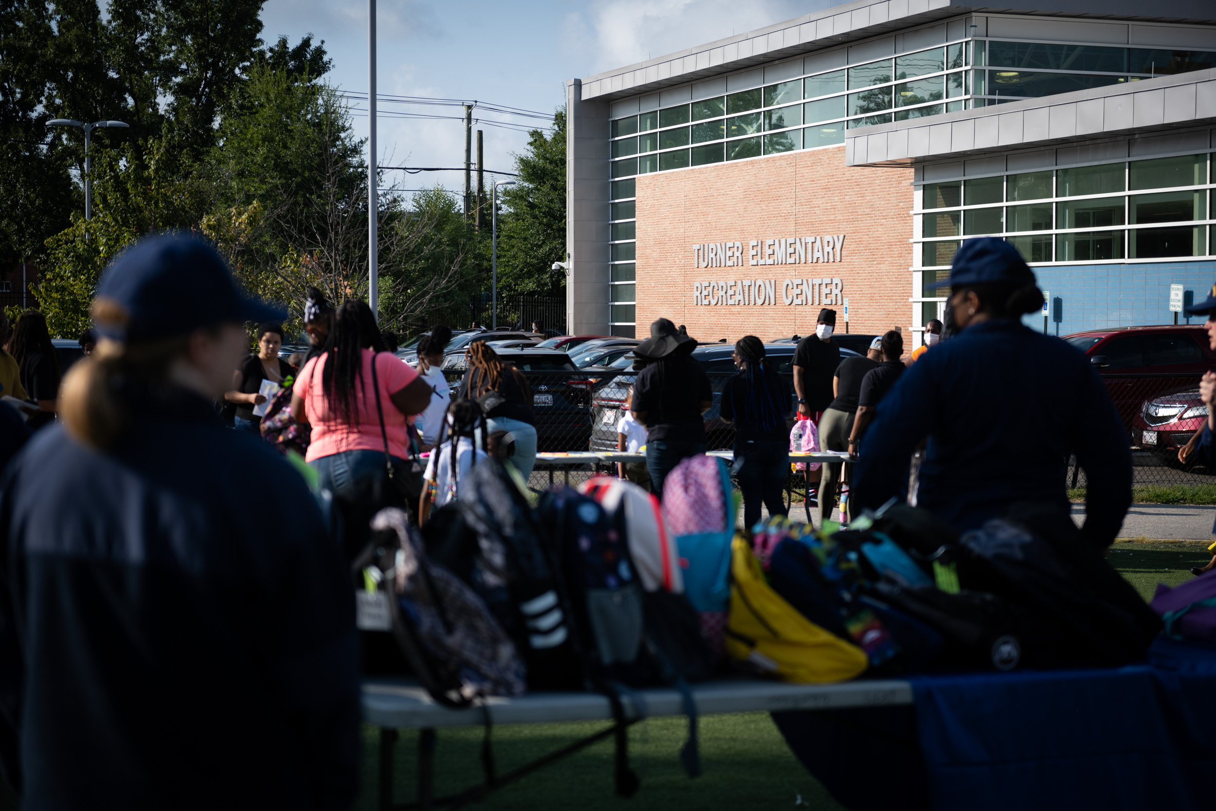 A crowd of adults and children gathers outdoors near tables covered with colorful backpacks.