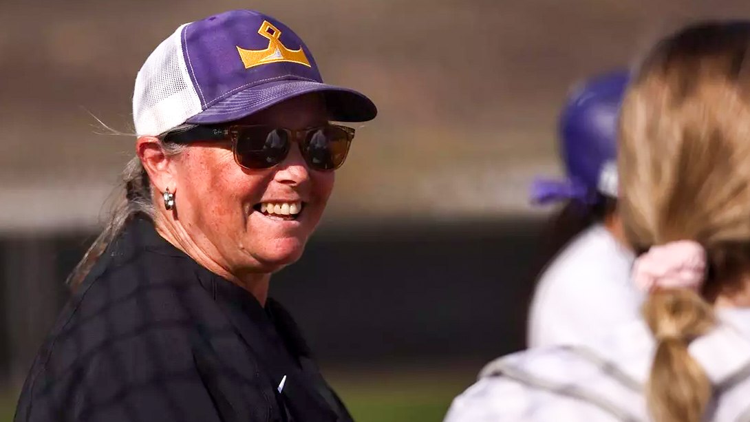 Smiling woman wearing a purple and gold baseball cap outdoors on an athletic field.