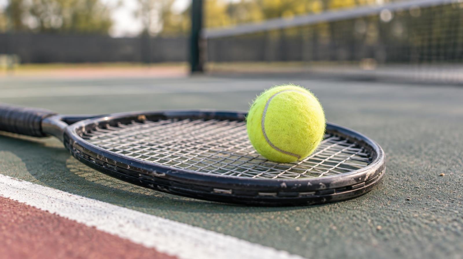 Yellow tennis ball resting on the strings of a tennis racket on a hard court.