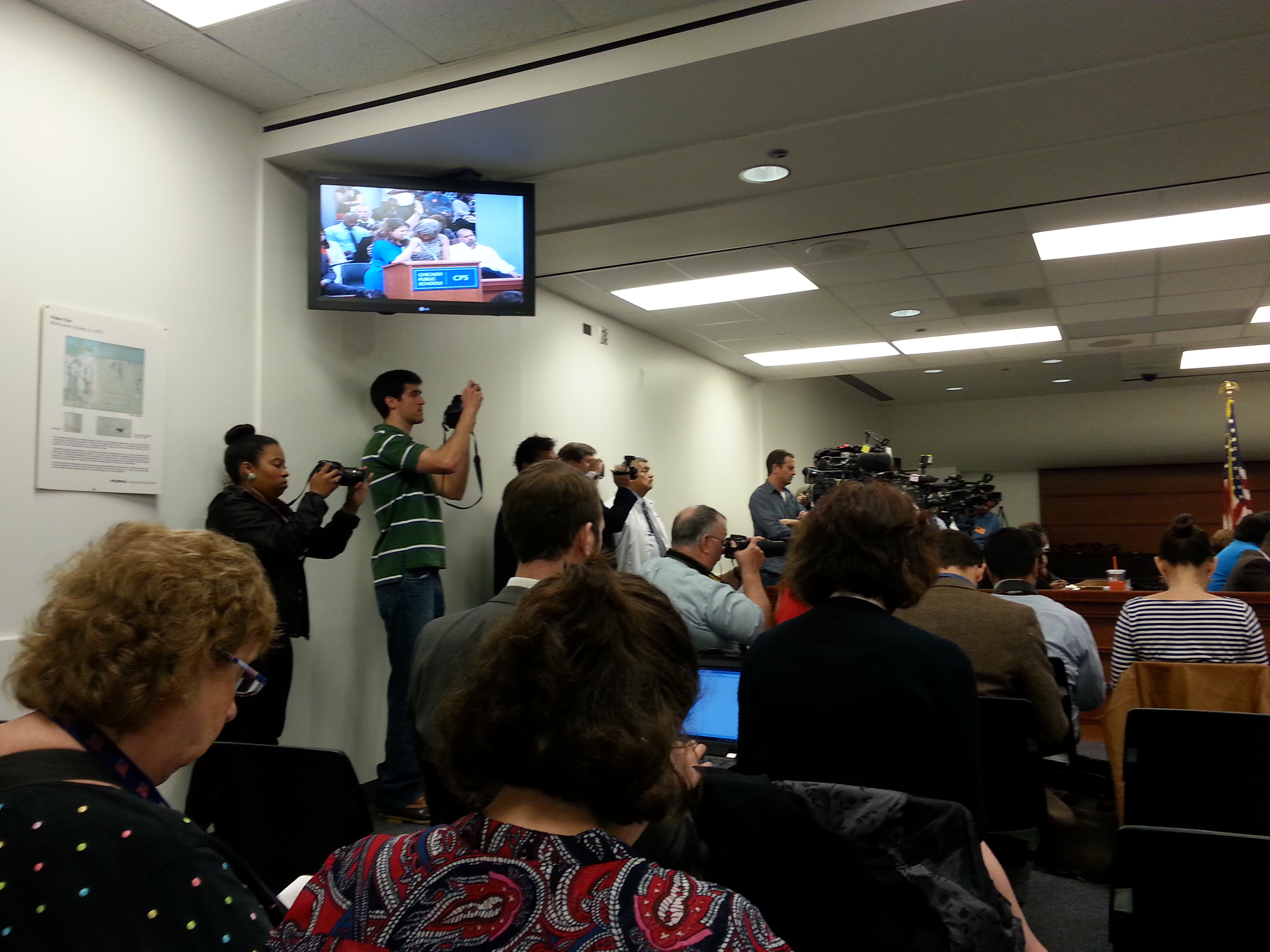 Crowded school board meeting room with audience members and journalists facing a dais.