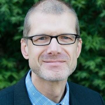 Man with shaved head and glasses wearing a dark blazer, photographed outdoors against green foliage.