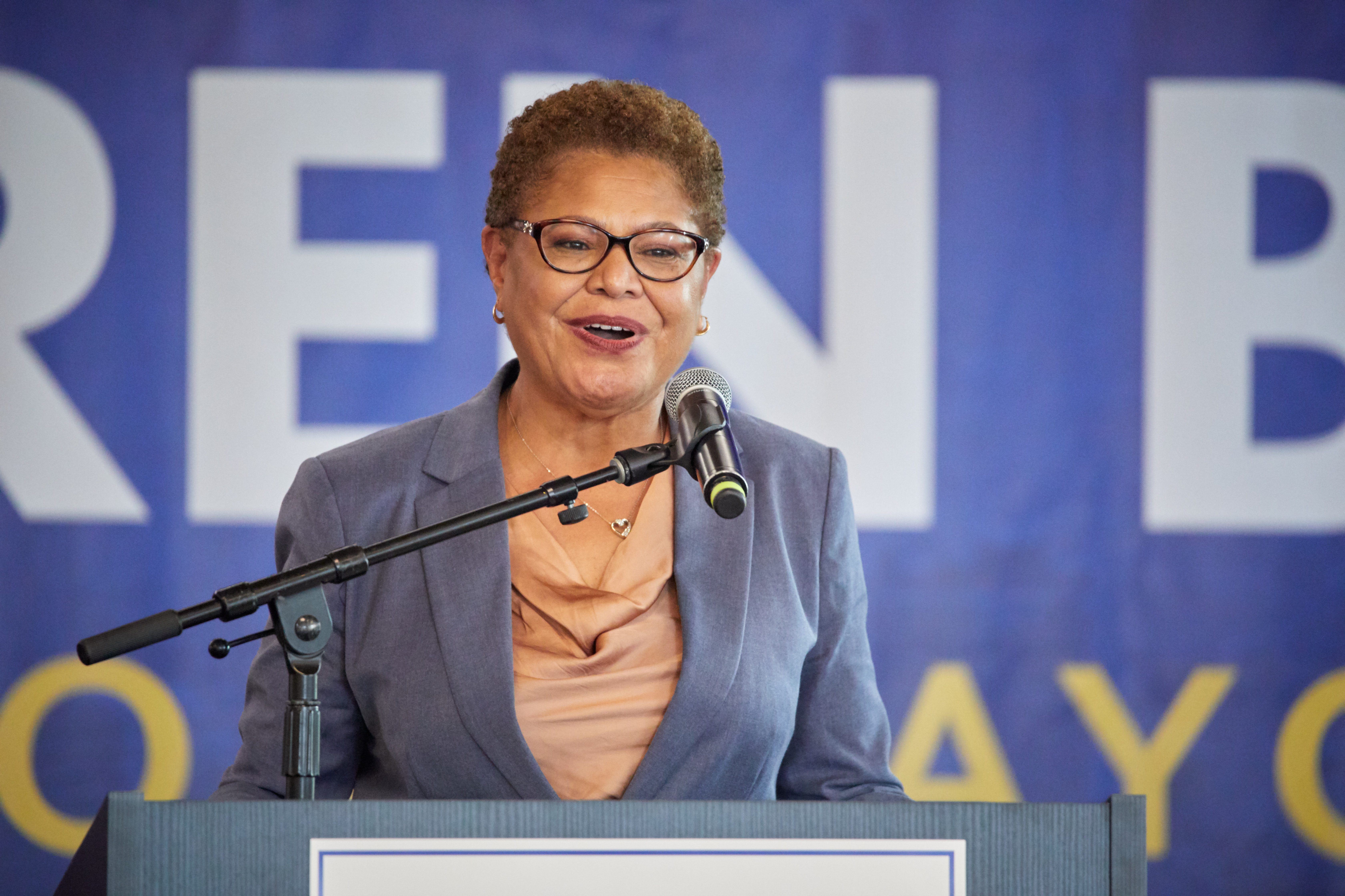 Woman speaking at a podium in front of a blue campaign banner reading Karen Bass for Mayor.