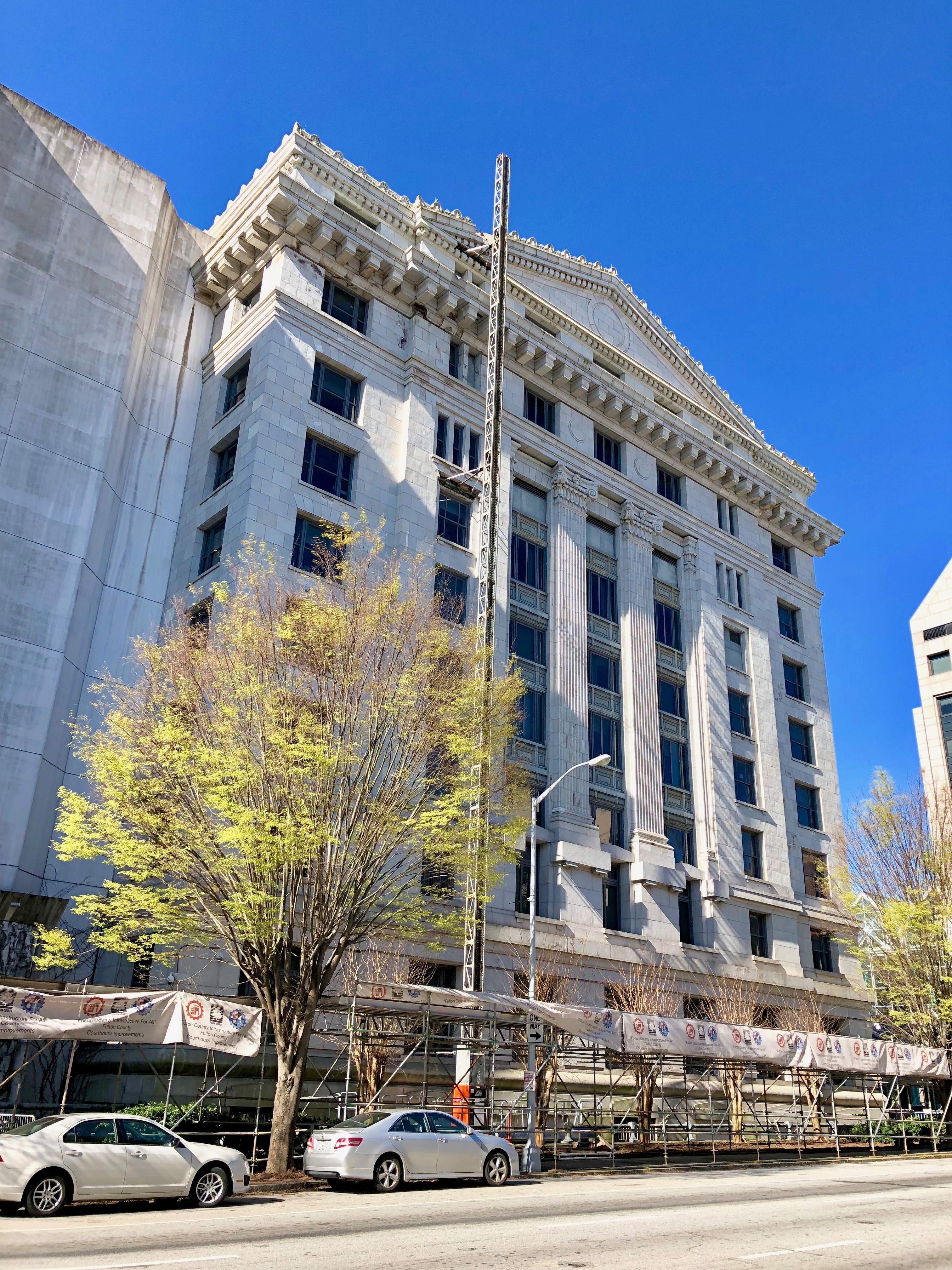 Neoclassical courthouse building with tall columns, scaffolding, and trees under a blue sky.