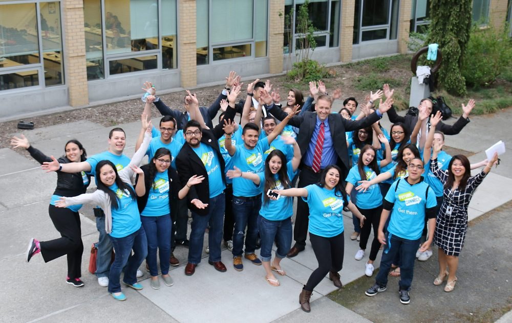 Large group of people in matching light-blue T-shirts posing outdoors with arms raised.