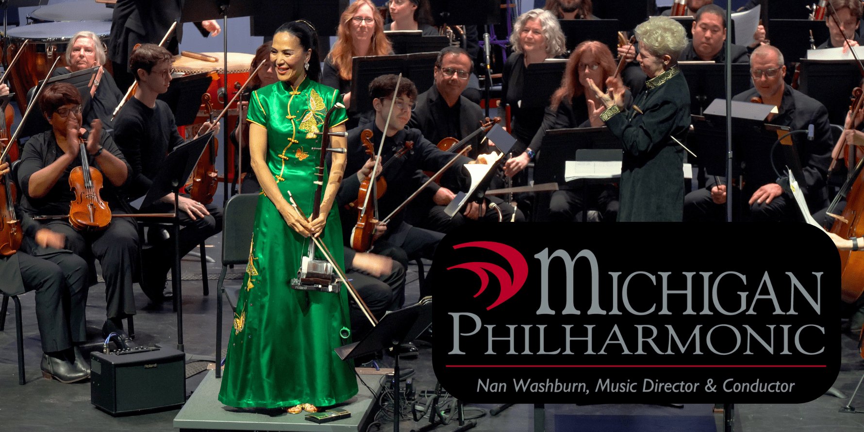 A conductor in a green dress holds a baton in front of a seated orchestra on stage.