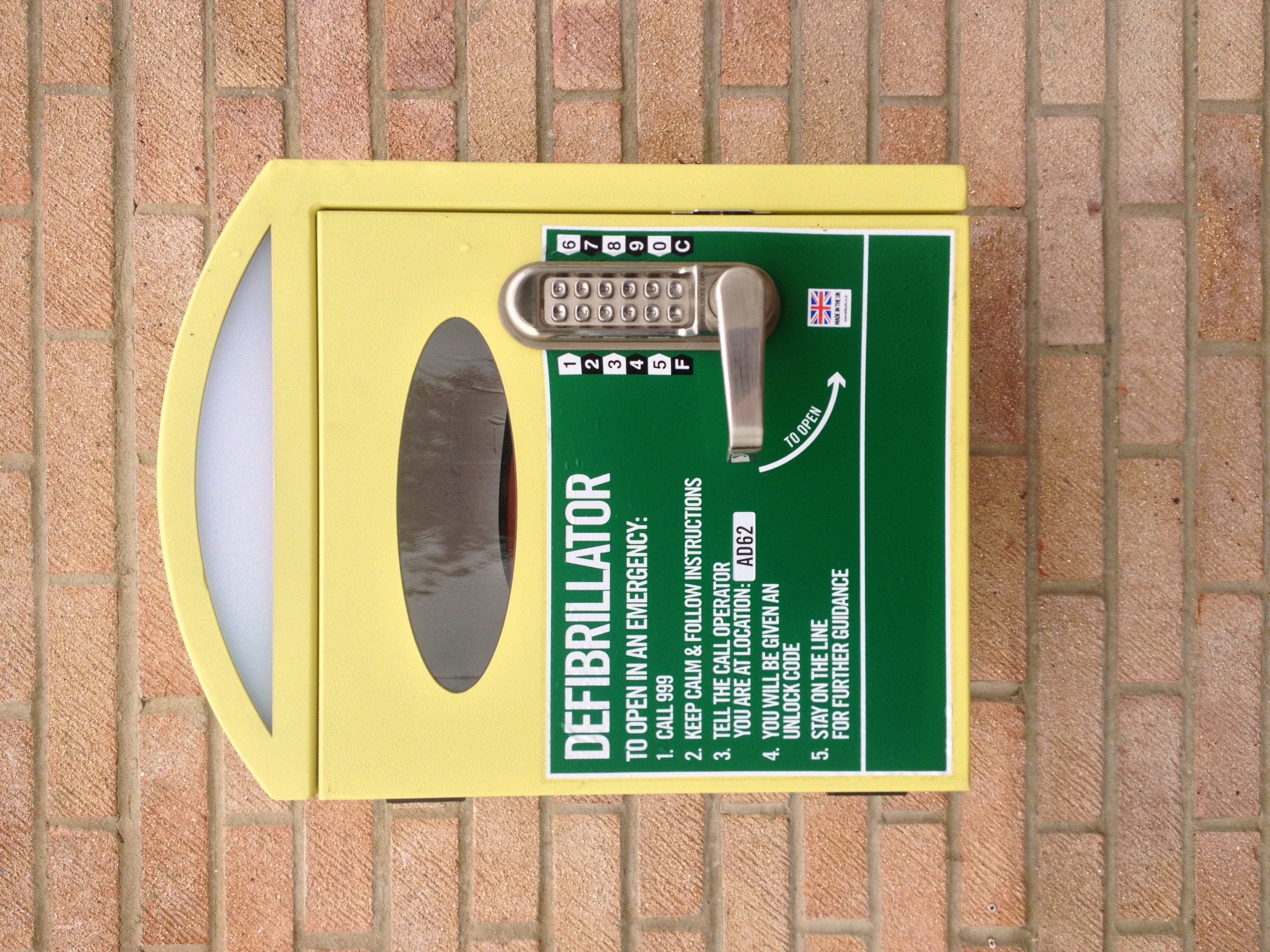 Yellow wall-mounted defibrillator cabinet with coded keypad lock on a brick wall.