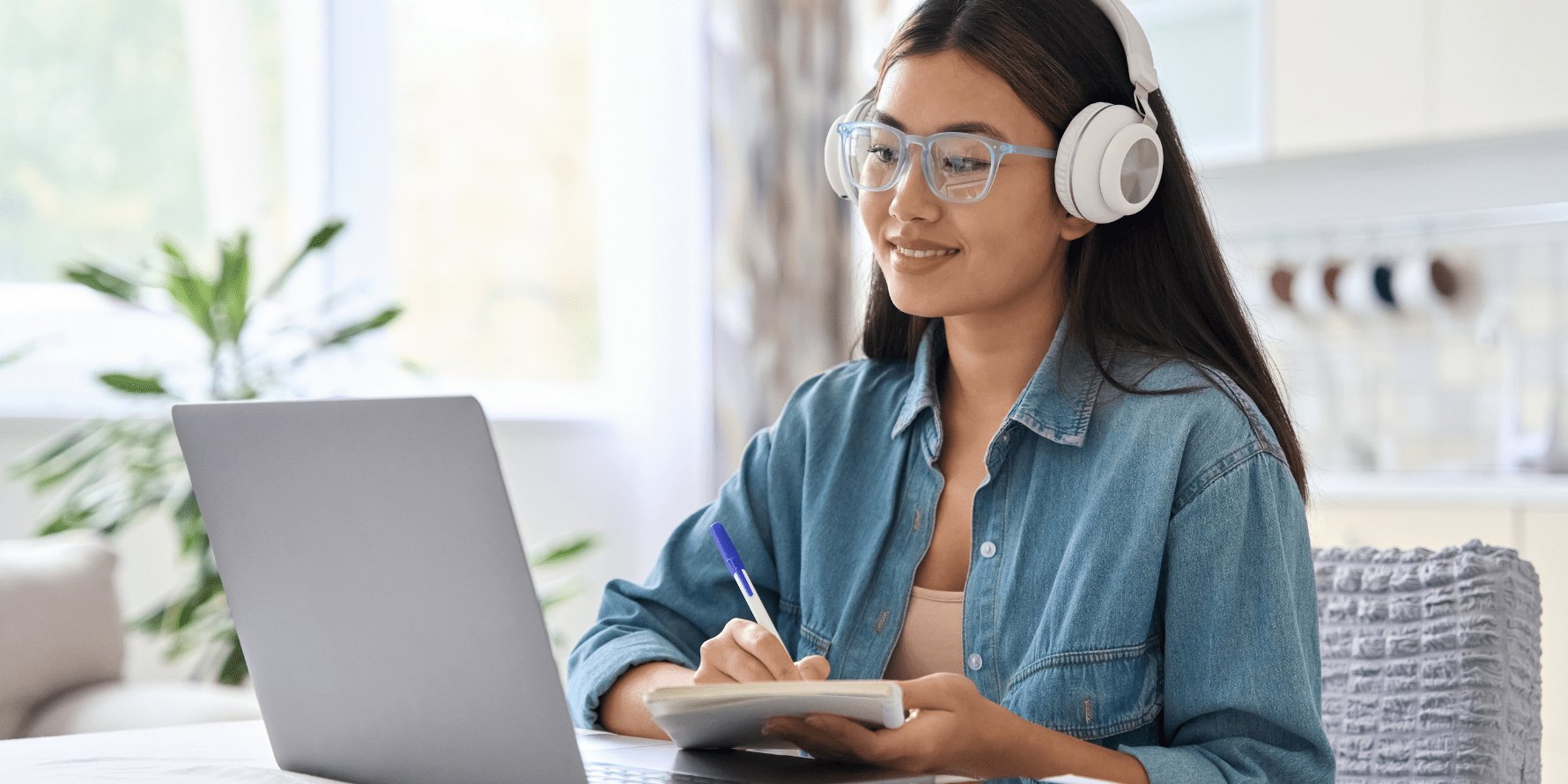 Young woman with headphones and glasses writing in a notebook while using a laptop.
