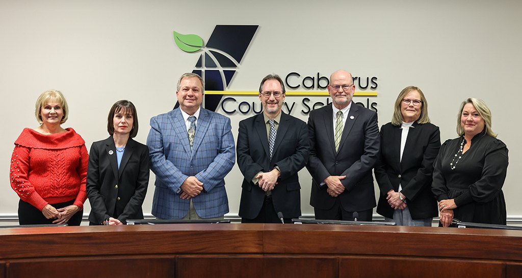 Seven adults in business attire standing in a row before a Cabarrus County Schools sign.
