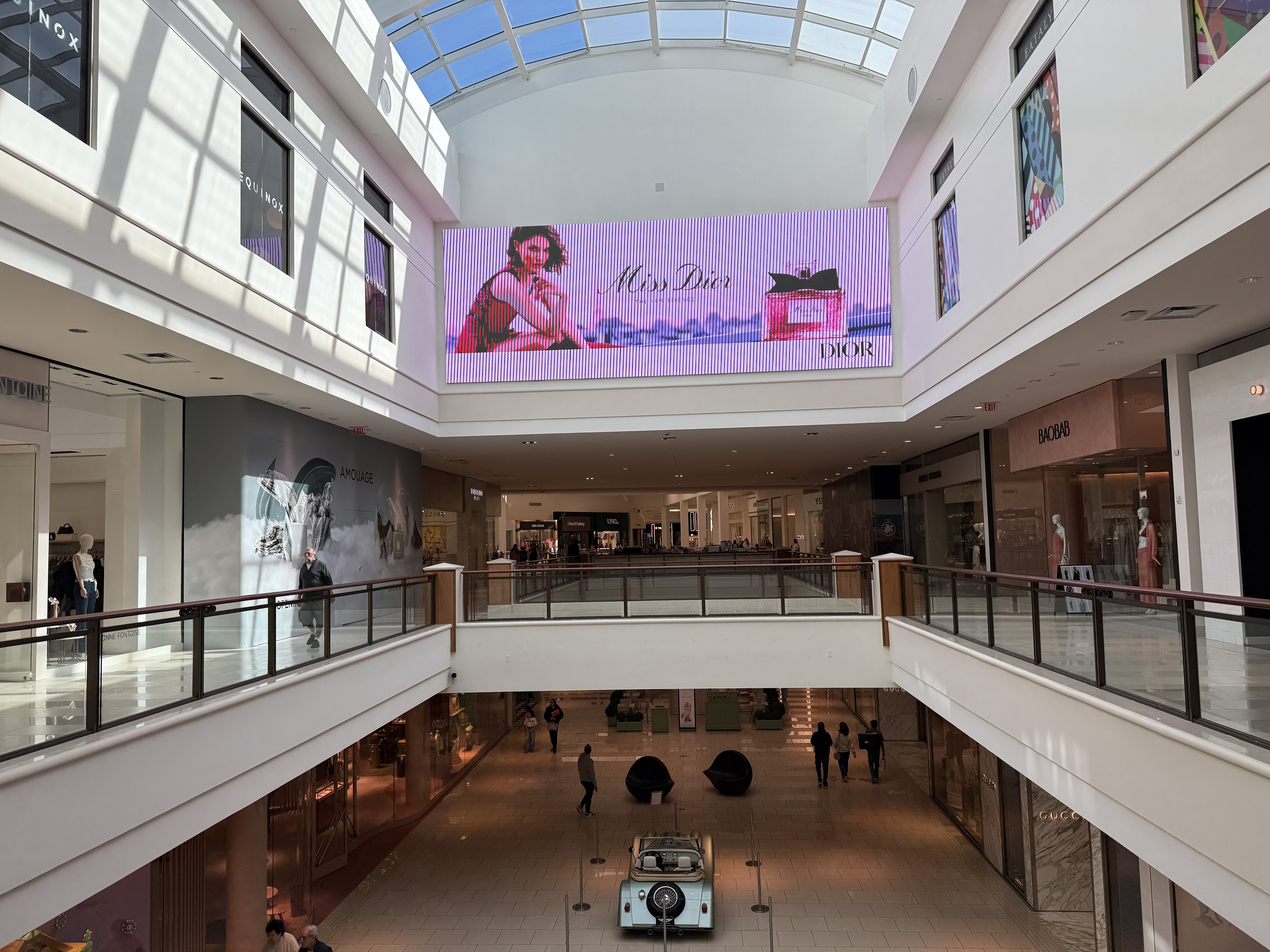 Two-story mall corridor with luxury storefronts and a large digital fragrance ad screen