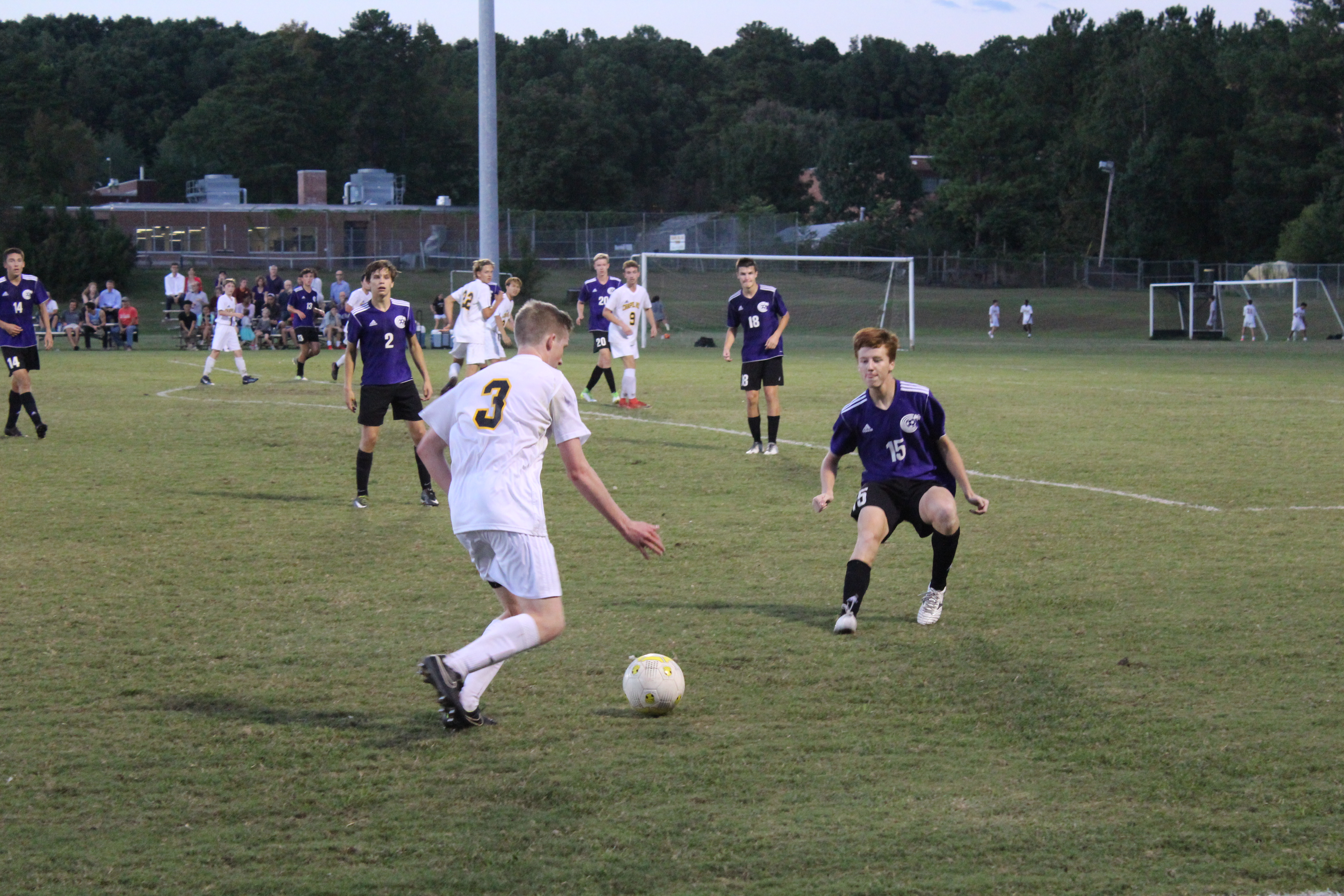 A player in a white jersey controls a soccer ball while a defender in purple closes in on a grass field.