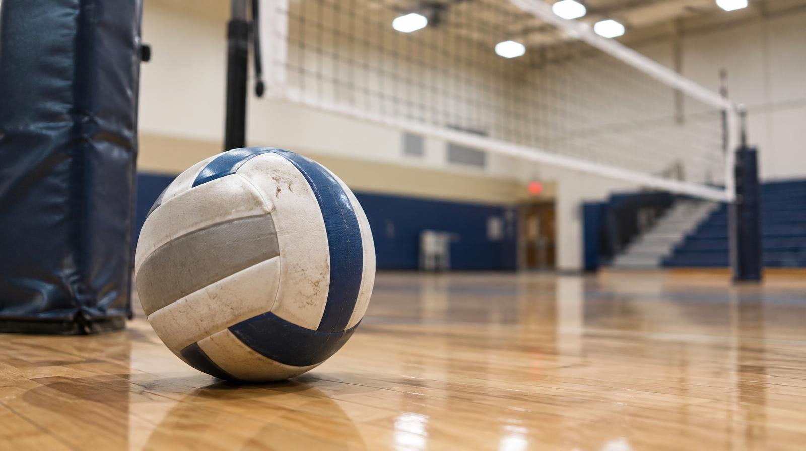 A volleyball resting on a hardwood gym floor near a volleyball net post.