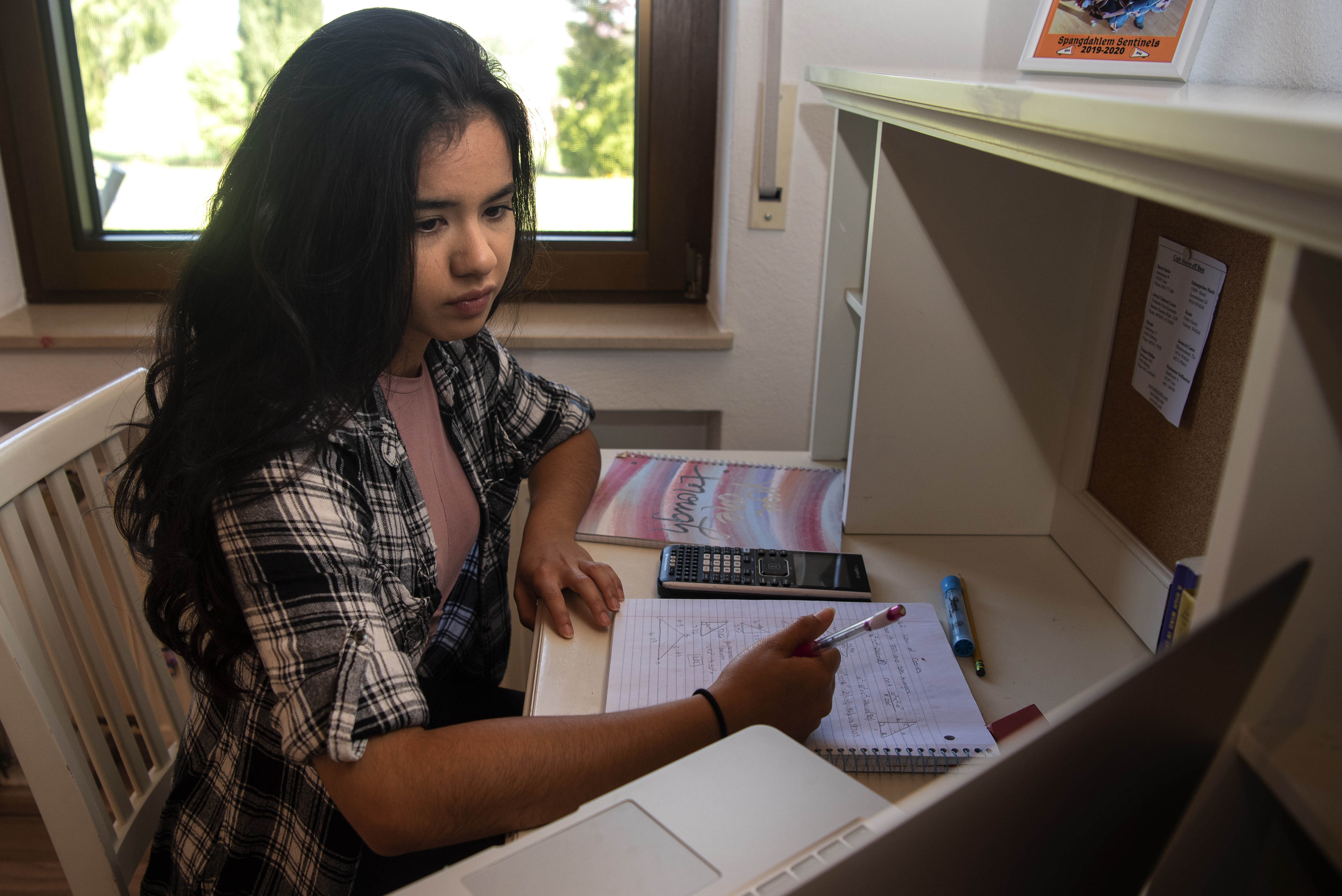 A teenage girl writes in a notebook at a desk with a calculator nearby.