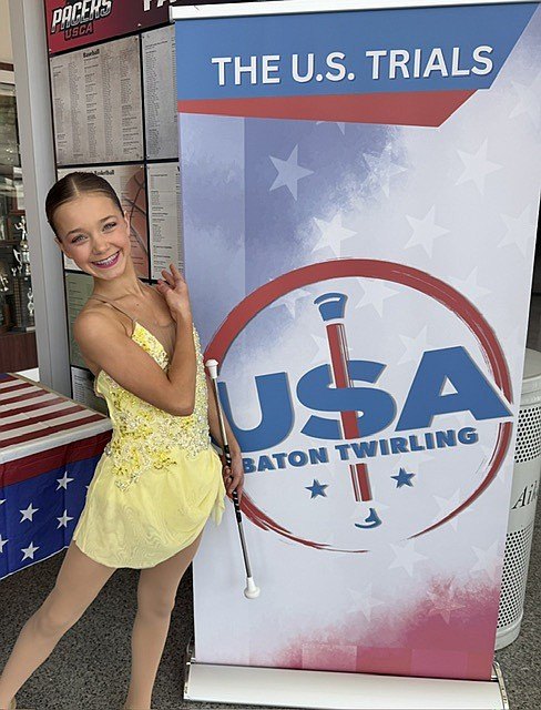 A smiling girl in a yellow sequined leotard holds a baton beside a competition banner.