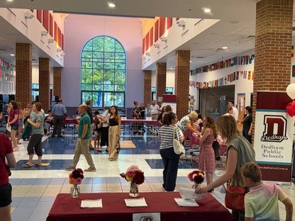 Crowd of adults in a school lobby decorated with international flags and a Dedham Public Schools banner.