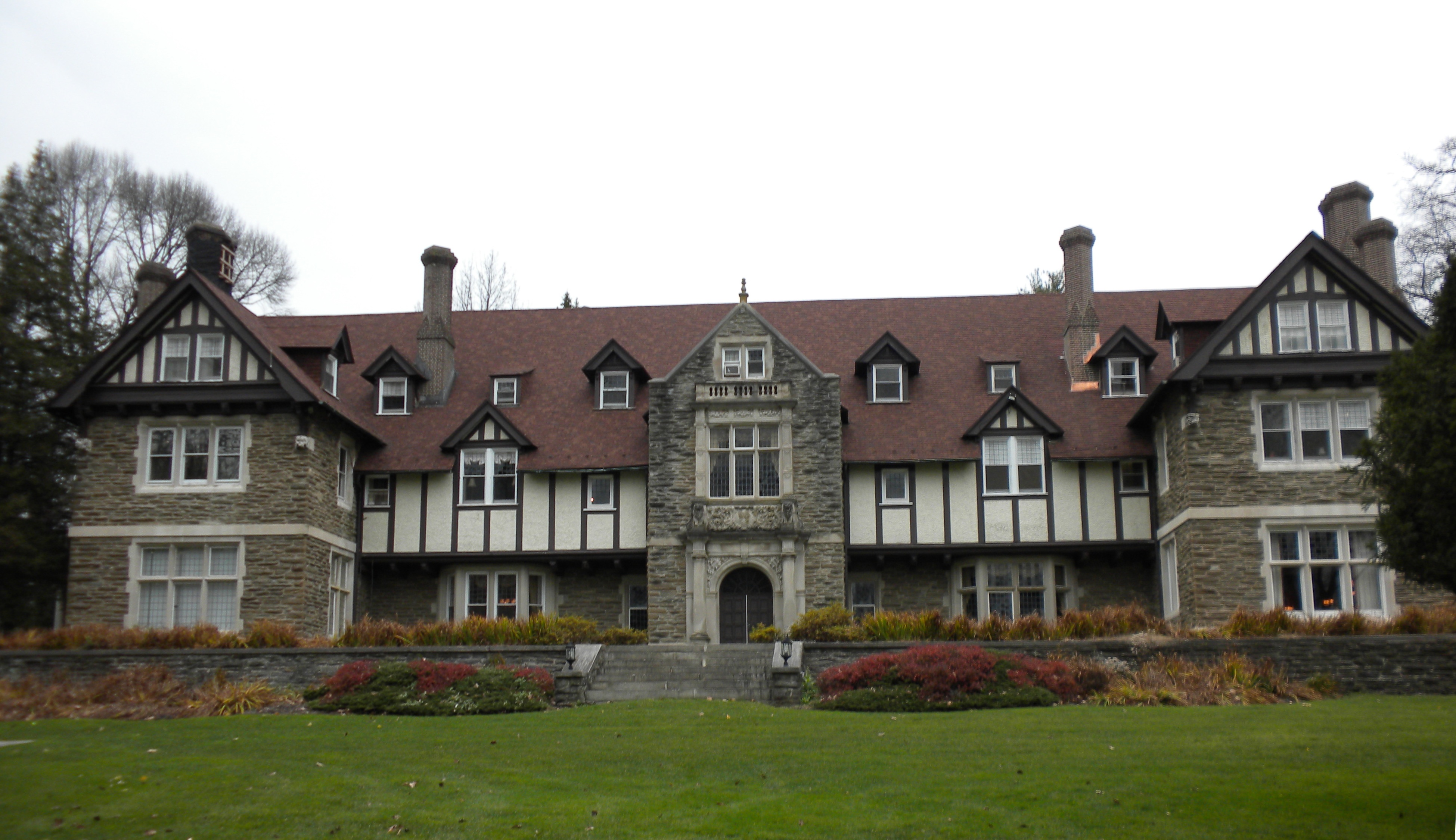 Large Tudor Revival stone mansion with red roof and chimneys on a manicured lawn.