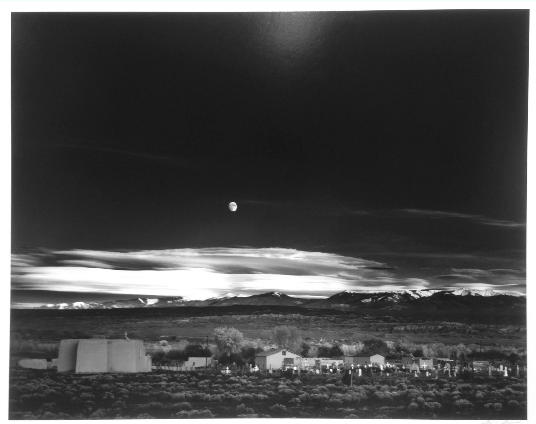 Black-and-white photo of a moonrise over adobe buildings with snow-capped mountains behind.