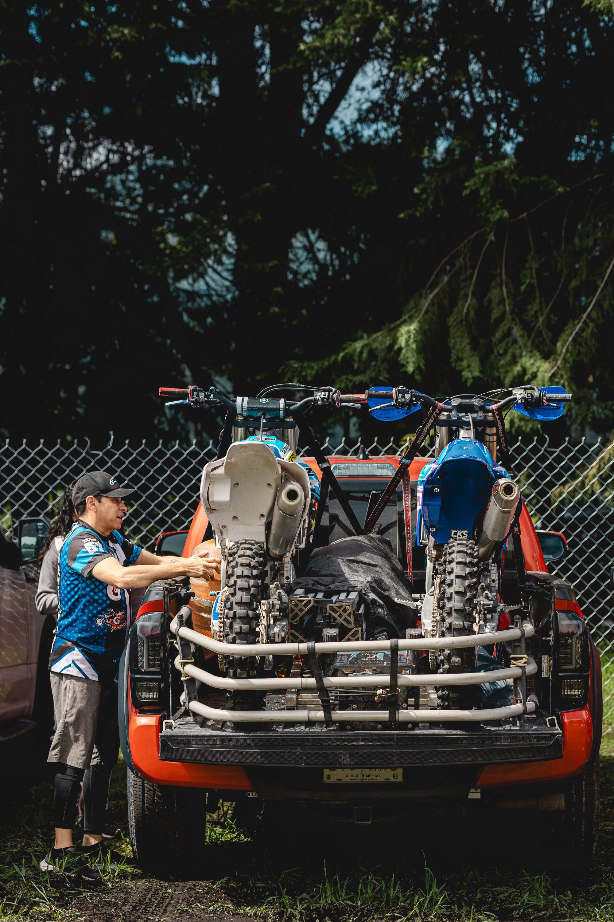 Two motocross bikes loaded on a pickup truck parked outdoors near a fence.