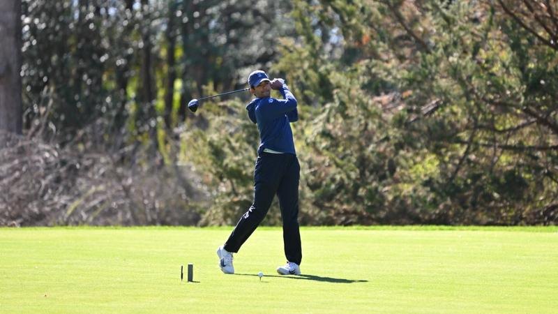 Golfer in blue shirt completes a full follow-through swing on a tree-lined course.
