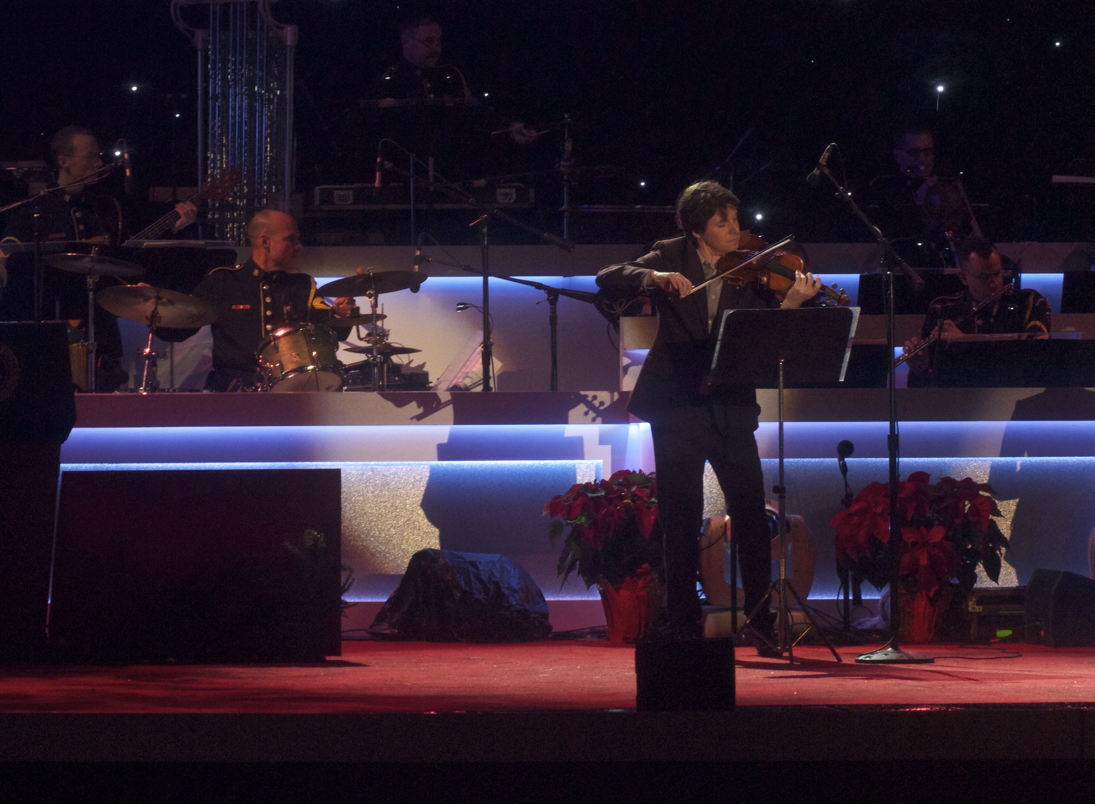 A violinist performs on a lit stage with an orchestra seated behind.