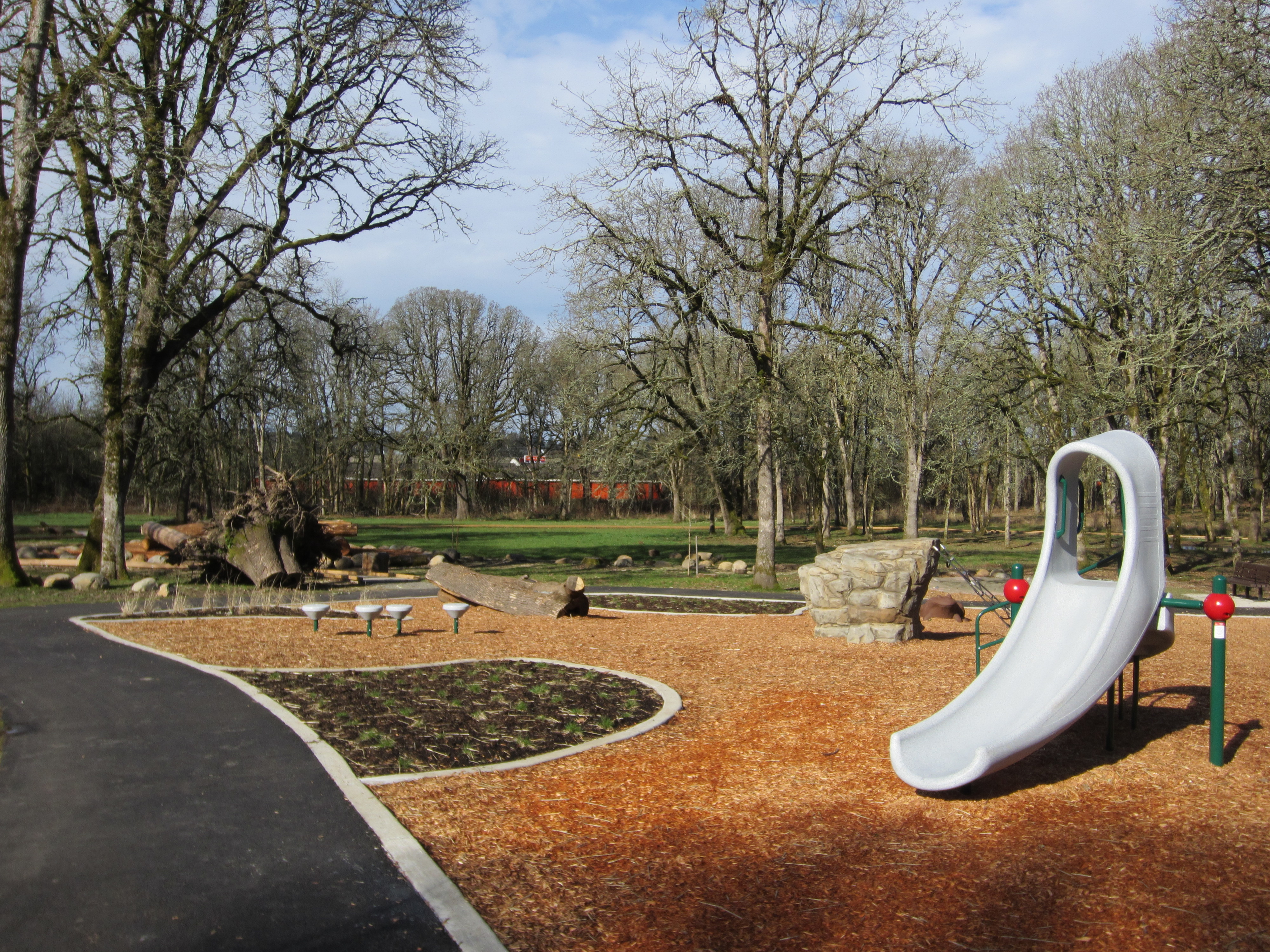 Playground with curved slide, natural rock and log elements, and wood chip surfacing.
