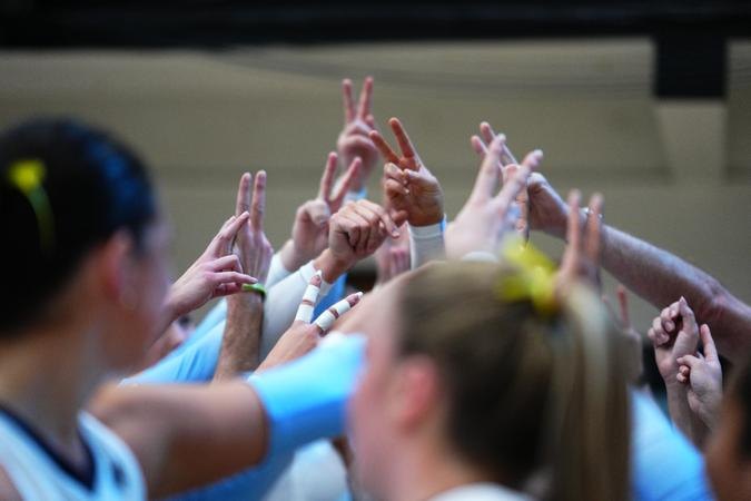 Female volleyball players in light blue uniforms form a team huddle, arms raised.