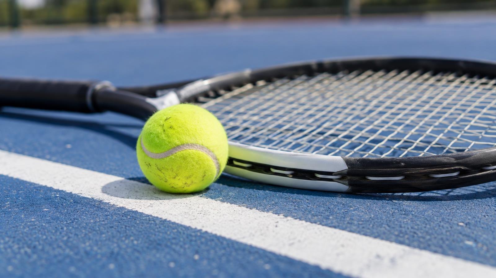 A tennis racket and yellow ball resting on a blue hard court near the white baseline.
