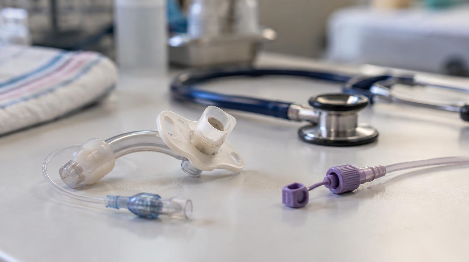 A tracheostomy tube and feeding tube connector beside a stethoscope on a clinical surface.