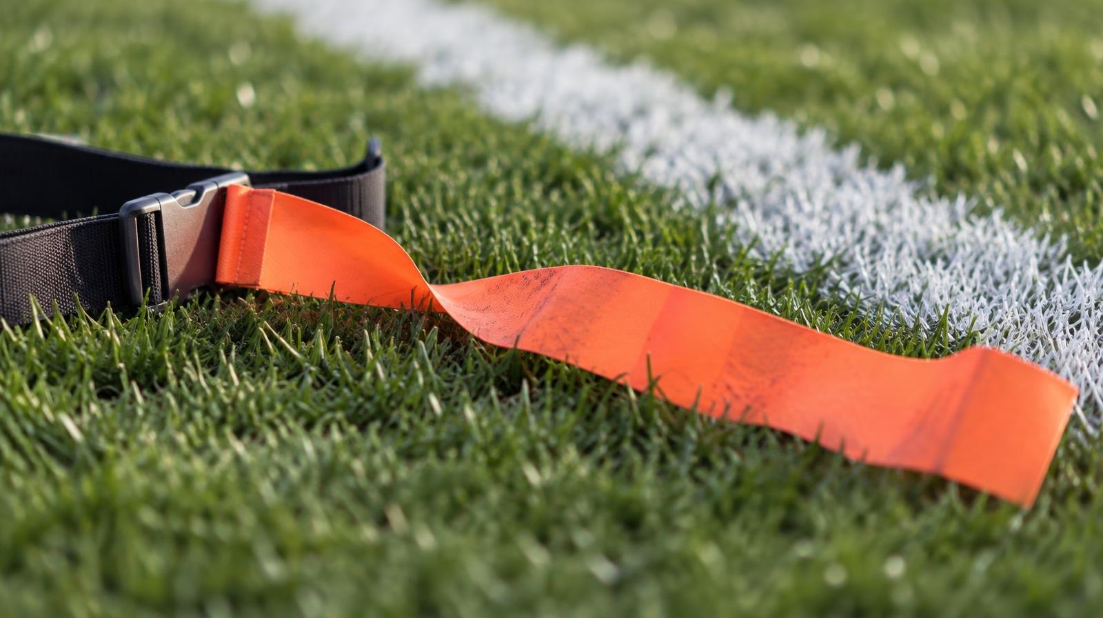 An orange flag football flag on a belt clip lying on green grass beside a white yard line.