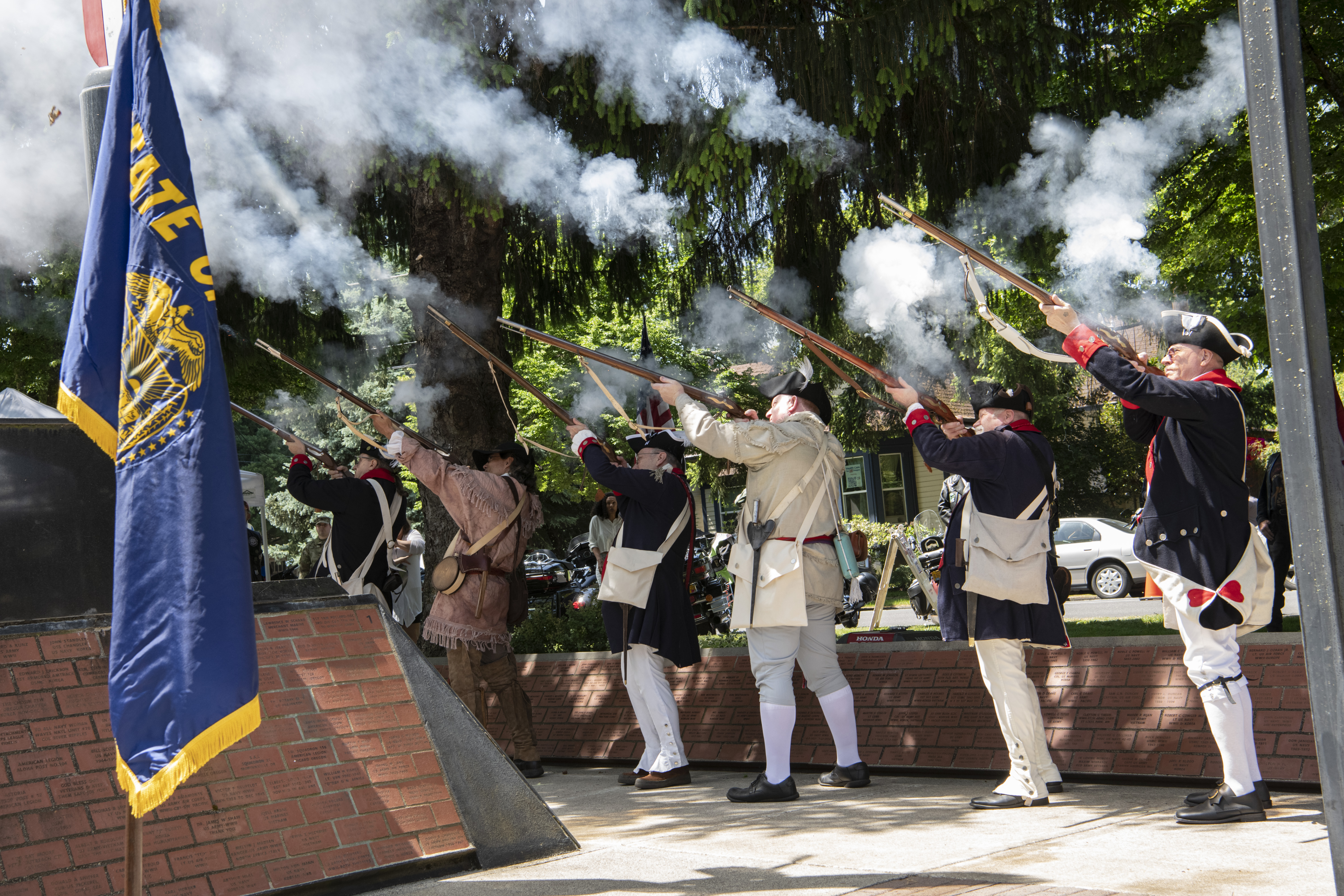Men in colonial-era uniforms fire muskets in a line at an outdoor ceremony, smoke rising.