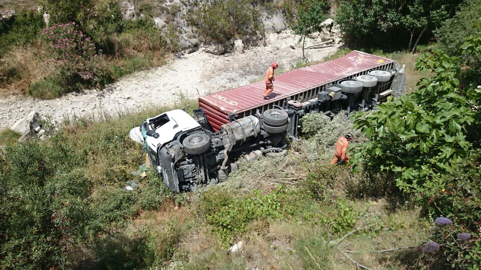 Overturned tractor-trailer on its side in brush with two safety workers nearby.
