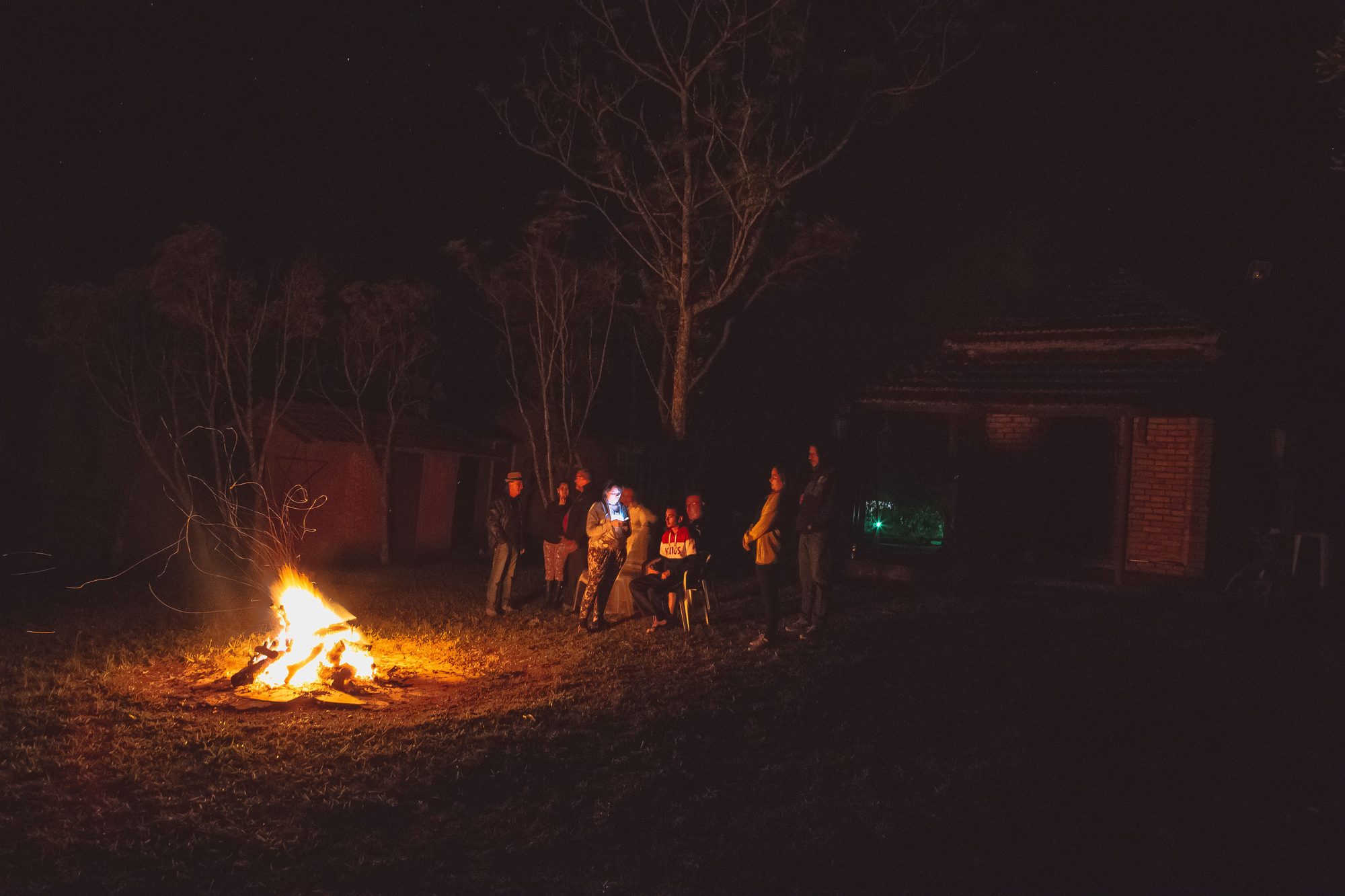 A group of people enjoy a cozy bonfire outdoors at night, surrounded by trees and a house.