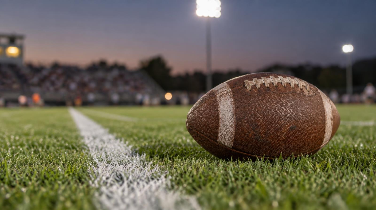 An American football resting on green grass beside a white yard line under bright stadium lights.