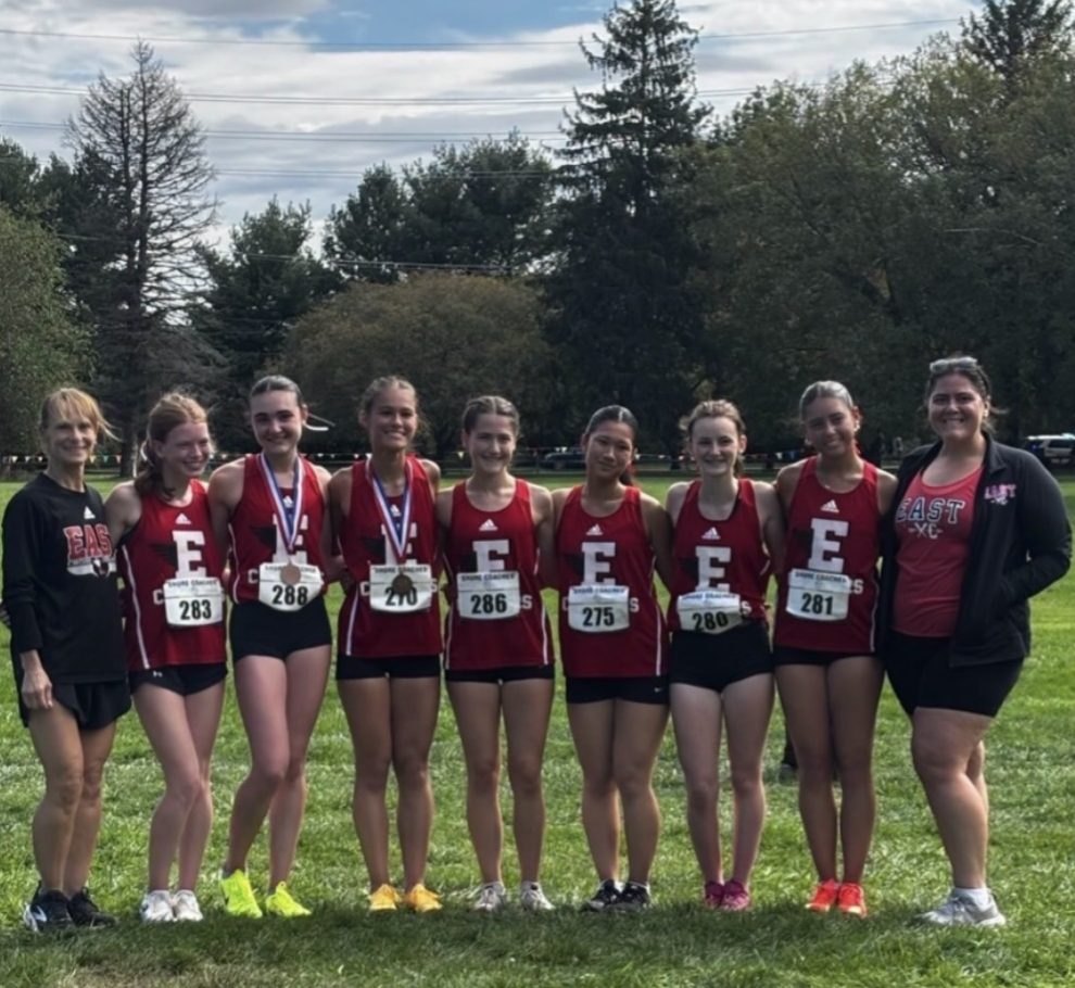 Seven young female runners in red uniforms with race bibs pose with two adults on grass.