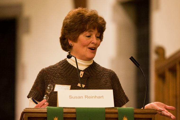 Woman at a wooden podium with a microphone moderating a candidate forum.